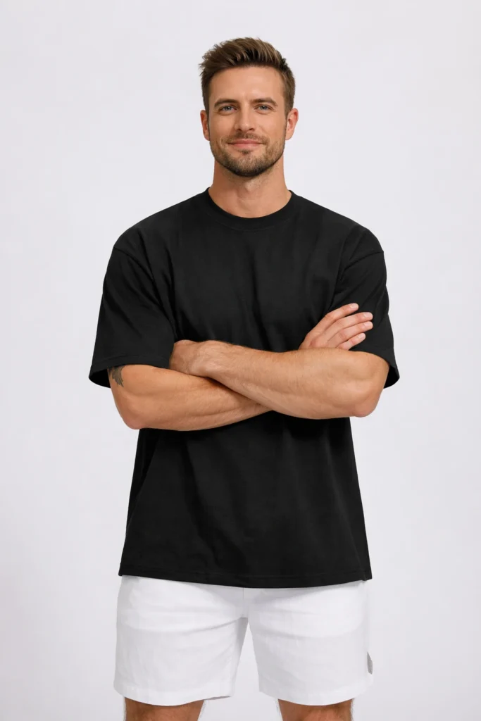 A studio portrait of a man wearing a black oversized T-shirt paired with white shorts. The relaxed fit, solid color, and simple styling create a modern, versatile look ideal for everyday wear and contemporary casual or streetwear collections.