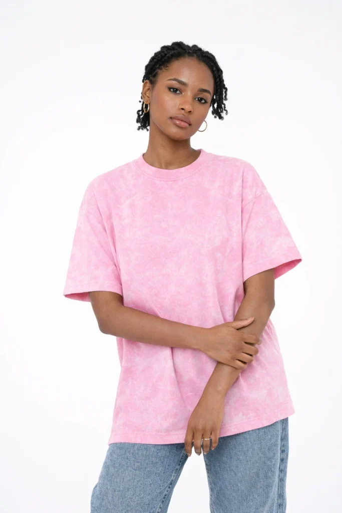 A clean studio portrait of a woman wearing a pink washed oversized T-shirt paired with light denim. The relaxed fit and subtle faded texture create a soft, modern look ideal for everyday wear and casual streetwear collections.