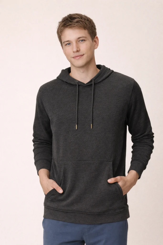 A studio lifestyle image featuring a male model wearing a heather charcoal pullover hoodie with a clean, lightweight construction. The hoodie includes a relaxed fit, soft knit fabric, adjustable drawstring hood with metal tips, ribbed cuffs and hem, and a front kangaroo pocket. The smooth texture and neutral tone make it suitable for modern casualwear, athleisure collections, and private label hoodie programs. Ideal for brands focusing on everyday comfort, minimalist design, and versatile seasonal layering.
