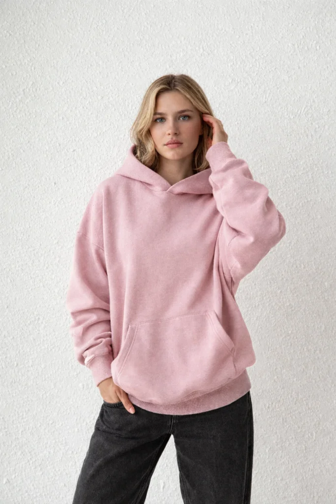 A clean lifestyle image featuring a women’s blush pink oversized pullover hoodie with a relaxed, modern silhouette. Made from soft fleece fabric, this hoodie includes dropped shoulders, ribbed cuffs and hem, and a classic kangaroo pocket for everyday comfort. The muted pink tone paired with a textured neutral wall creates a minimalist aesthetic, making it ideal for casual wear, cozy loungewear, and private label hoodie collections focused on comfort, versatility, and contemporary streetwear style.