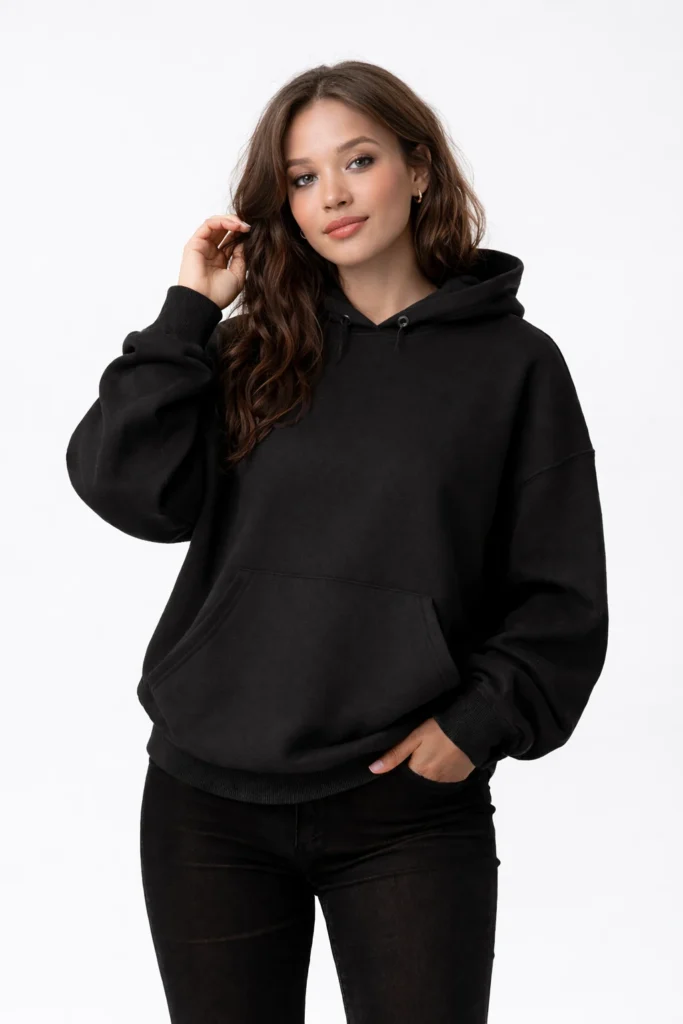 A clean studio lifestyle image featuring a women’s black oversized pullover hoodie with a relaxed, modern silhouette. Made from soft fleece fabric, the hoodie includes a classic hood, ribbed cuffs and hem, and a spacious kangaroo pocket for everyday comfort. The minimalist design and neutral background emphasize versatility, making this style ideal for casual wear, streetwear brands, and private label hoodie collections focused on comfort, simplicity, and contemporary fashion.