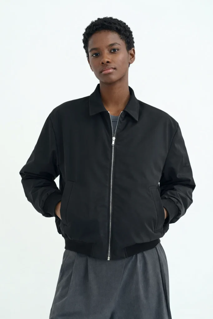 A clean studio image featuring a woman wearing a classic black bomber jacket with a relaxed, slightly oversized fit. Designed with a smooth matte finish, full front zipper, ribbed cuffs, and a minimalist collar, this jacket delivers a timeless and versatile silhouette. Paired with neutral-toned trousers, the look emphasizes everyday wearability and modern simplicity, making it ideal for women’s casual outerwear collections, private label bomber jackets, and OEM/ODM fashion brands targeting premium minimal and urban markets.
