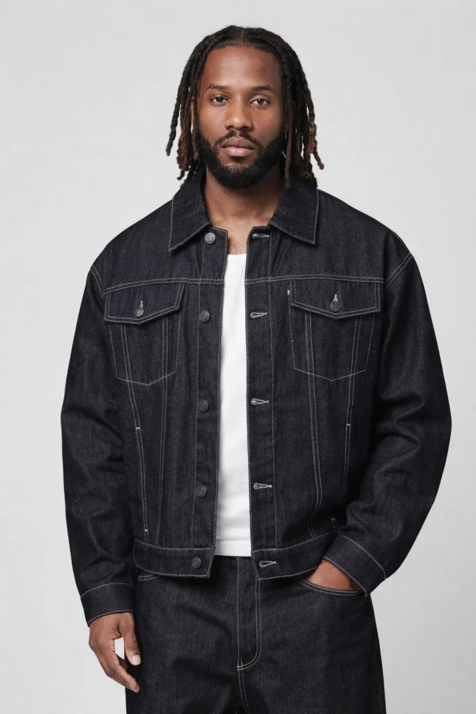 A clean studio image featuring a men’s raw black denim jacket designed with a classic trucker structure and a slightly relaxed fit. Crafted from heavyweight denim, this jacket showcases contrast stitching, metal button closures, and symmetrical chest flap pockets for a bold, durable look. Styled with a white T-shirt and matching dark denim bottoms, it delivers a strong workwear-inspired aesthetic with modern streetwear appeal. Ideal for OEM / private label denim programs focused on premium casualwear and timeless core styles.