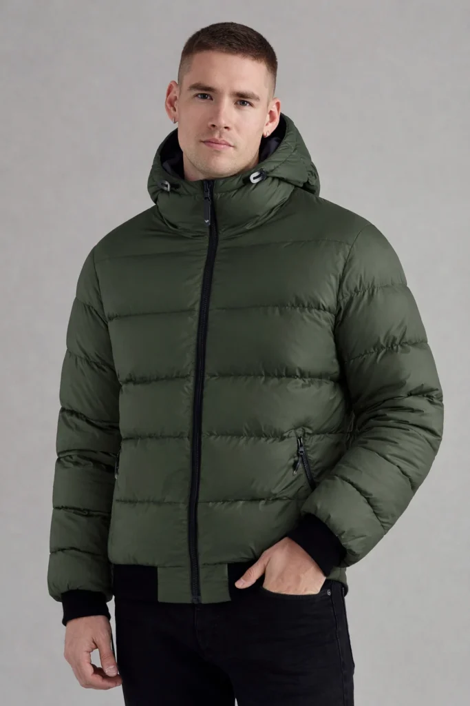 A clean studio shot featuring a men’s hooded puffer jacket in a deep olive green tone. Designed with horizontal quilted panels, a full front zipper, and an adjustable padded hood, this jacket delivers reliable warmth while maintaining a streamlined, modern silhouette. Ribbed cuffs and zippered side pockets enhance practicality for everyday wear. Ideal for autumn and winter collections, this style suits OEM / private label outerwear programs focused on functional, urban, and outdoor-inspired men’s apparel.