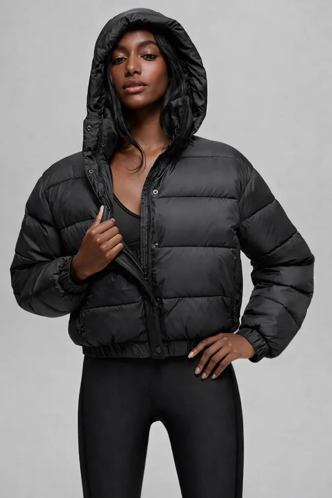A clean studio image showcasing a women’s black hooded puffer jacket with a cropped silhouette and horizontal quilted padding. The jacket features a full-zip front with snap-button placket, elastic cuffs, and a structured insulated hood for added warmth and protection. Designed for a sleek, modern fit, this style blends performance outerwear with contemporary athleisure aesthetics—ideal for OEM and private label winter collections targeting urban, casual, and fashion-forward womenswear markets.