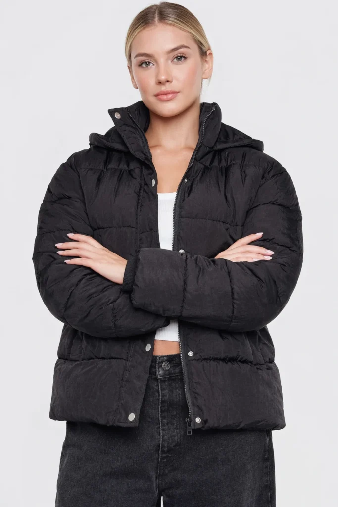 A studio-style image featuring a women’s black quilted puffer jacket with a relaxed, modern silhouette. The jacket is designed with horizontal quilting, a full-zip front combined with snap-button closure, and a lightly padded structure for everyday warmth without bulk. Worn over a cropped top and high-waisted denim, this versatile outerwear piece delivers a clean, contemporary look ideal for casual wear, streetwear collections, and OEM or private label winter apparel lines focused on minimalist, urban styling.