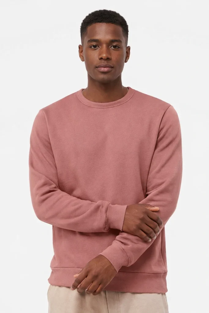 A studio portrait featuring a men’s dusty rose crewneck sweatshirt with a clean, minimalist design. Made with soft cotton fleece fabric, the sweatshirt includes a classic ribbed neckline, cuffs, and hem for a structured yet relaxed fit. Styled with neutral-toned pants, this piece highlights a modern casual aesthetic ideal for everyday wear, streetwear collections, and OEM or private label apparel focused on comfort, simplicity, and versatile color palettes.