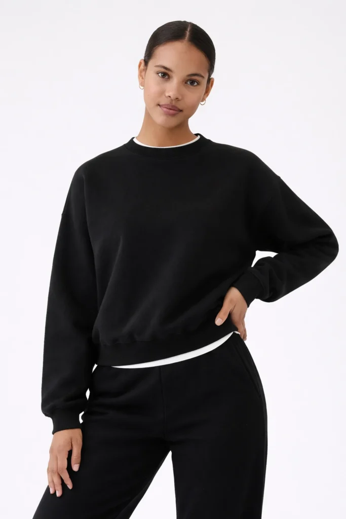 A clean, modern studio image showcasing a women’s black crewneck sweatshirt styled with matching sweatpants for a cohesive minimalist look. The sweatshirt features a relaxed fit, classic round neckline, and ribbed cuffs and hem, emphasizing comfort, versatility, and timeless appeal. Layered subtly over a white inner top, this look highlights everyday wearability and premium simplicity—ideal for custom loungewear collections, private label basics, and contemporary casual brands focused on elevated essentials.