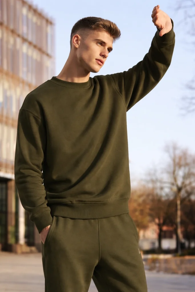 A contemporary outdoor lifestyle shot featuring a men’s olive green crewneck sweatshirt paired with matching jogger-style sweatpants. The sweatshirt showcases a clean, minimalist design with a classic round neckline, ribbed cuffs, and a relaxed yet structured fit. Styled in a natural urban setting with soft daylight, the look highlights versatility, comfort, and modern masculinity—ideal for premium athleisure collections, custom streetwear programs, and private label casualwear focused on everyday wear, neutral color palettes, and elevated basics.
