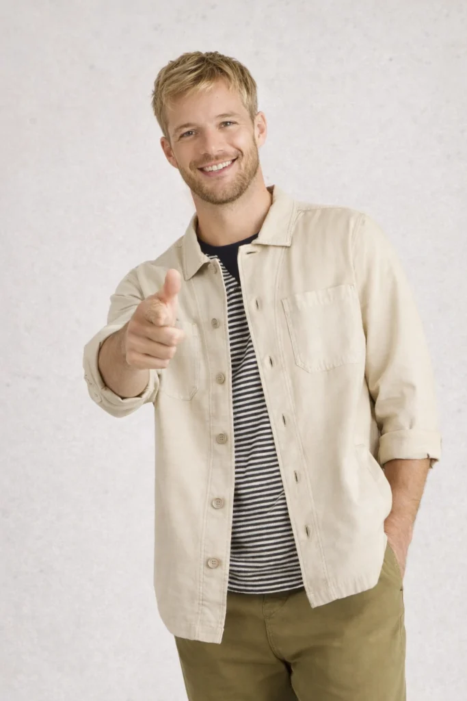 A bright studio-style image featuring a men’s beige overshirt jacket layered over a striped crewneck T-shirt and paired with olive green pants. The lightweight button-up overshirt offers a relaxed yet polished fit, making it ideal for casual city wear, transitional seasons, and modern lifestyle collections. Clean stitching, neutral tones, and an easy layering silhouette make this style perfect for private label, OEM/ODM apparel brands focused on versatile menswear essentials and everyday comfort.